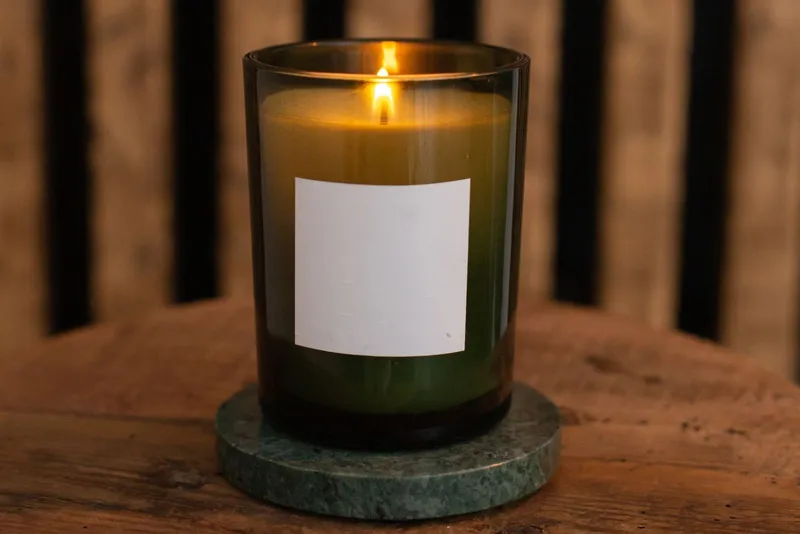 Bougie allumée dans un verre vert fumé à étiquette vierge, posée sur un dessous de verre en marbre vert sur une table en bois
