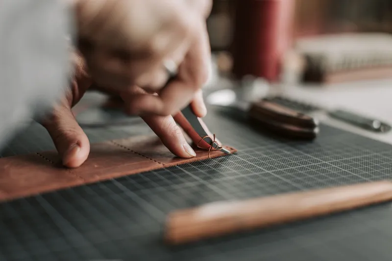 Mains d'artisan travaillant une bande de cuir fauve piquée, sur un tapis de coupe quadrillé