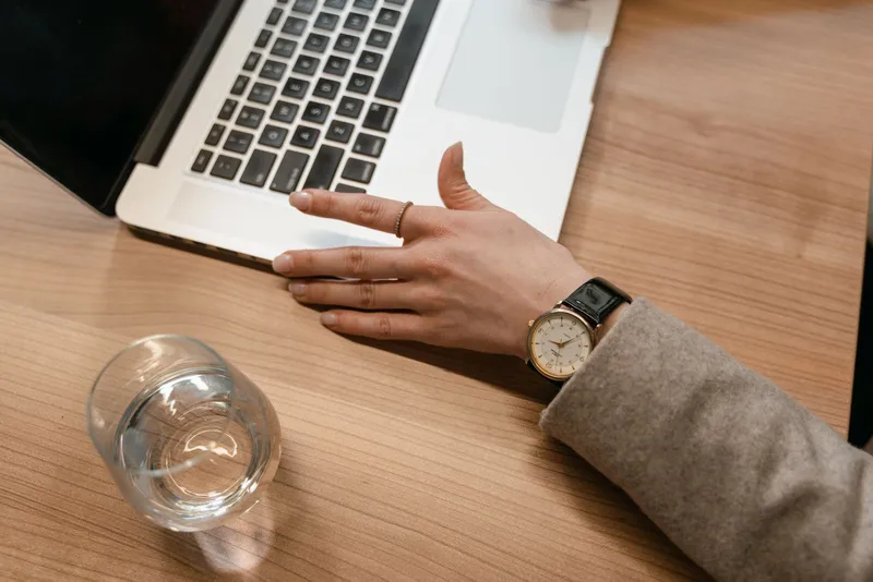 Poignet de femme portant une montre à cadran crème, boîtier doré et bracelet de cuir noir, posé sur un bureau en bois près d'un ordinateur portable